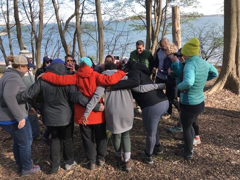 A group of adults are gathered in a circle outdoors, with their arms around each other. They appear to be in a wooded area near a body of water. The atmosphere seems to be one of camaraderie and connection, possibly during a team-building activity or a casual social gathering. The varying attire suggests a relaxed, informal setting.